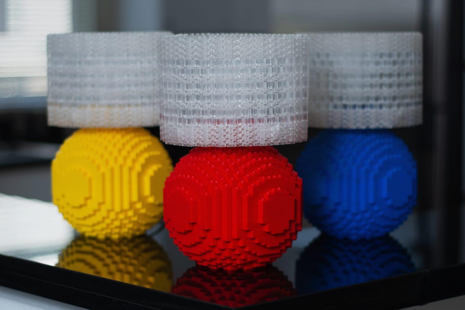 Three Splash of Color lamps with yellow, red, and blue brick bases topped with translucent brick shades, reflected on a glossy black surface.