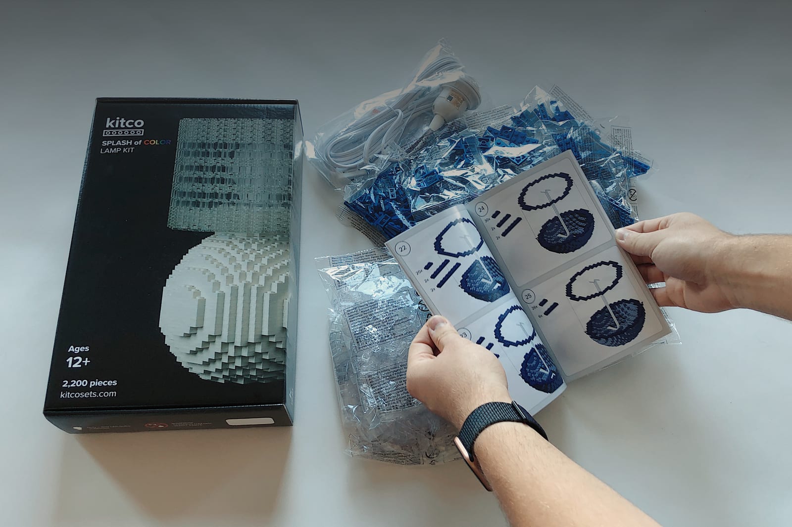 The Splash of Color Lamp Kit box beside bagged bricks, a white power cord, and hands holding the printed instruction booklet.