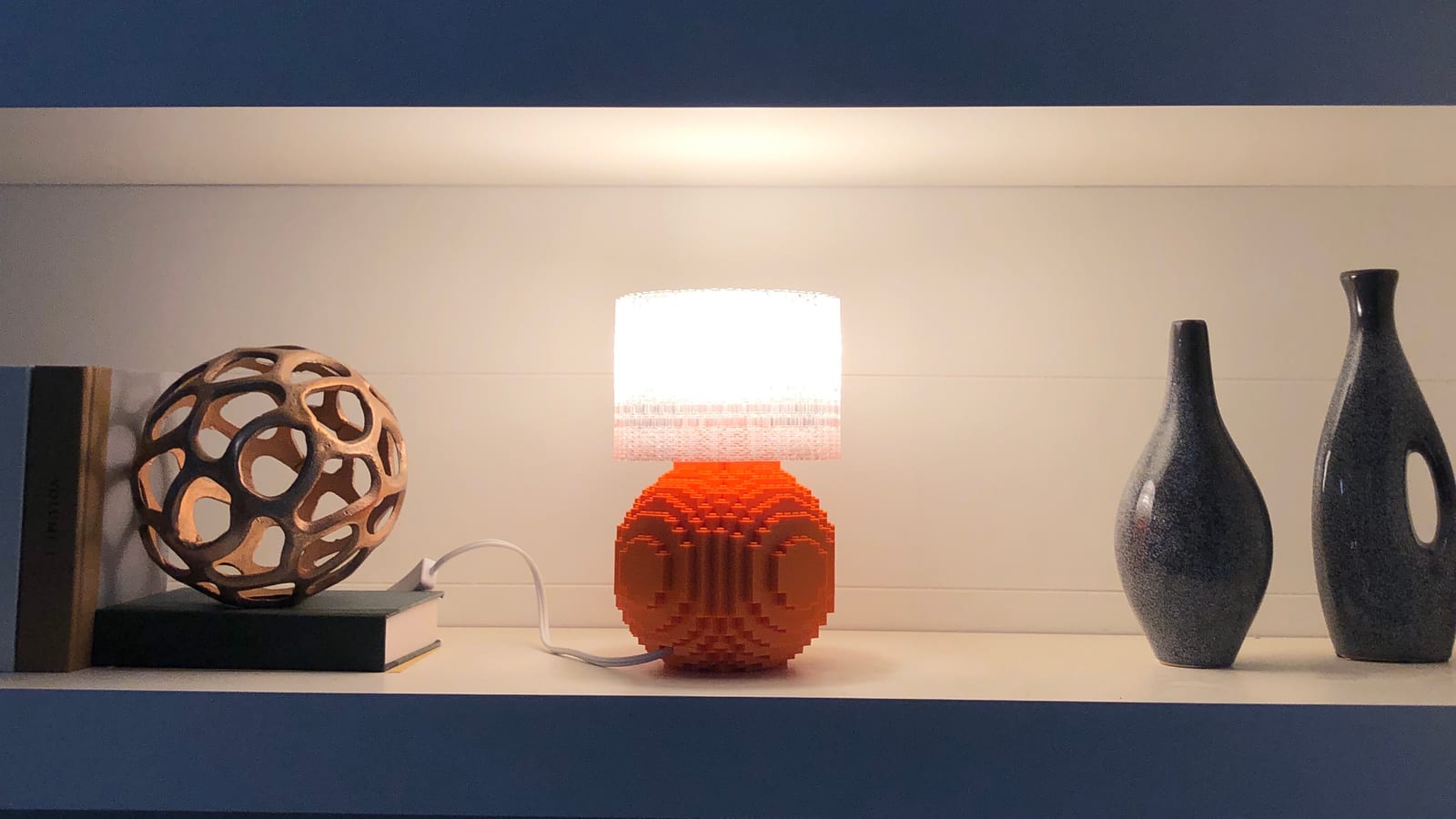 An orange Splash of Color lamp glowing on a styled shelf beside a wooden decorative sphere and two ceramic vases.